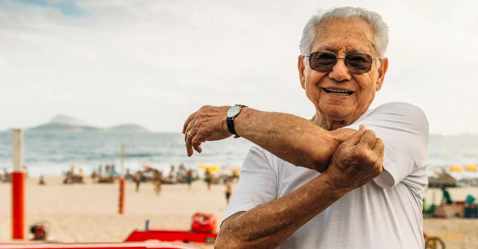 Idoso na orla de uma praia no rio de janeiro. Ele está de óculos e alongando o braço esquerdo.